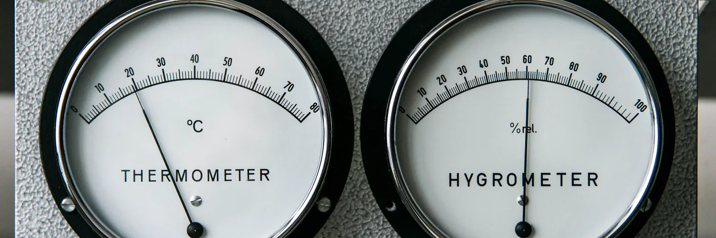 A wall-mounted thermometer and hygrometer, the two dials that describe what a heat cabinet is actually doing.