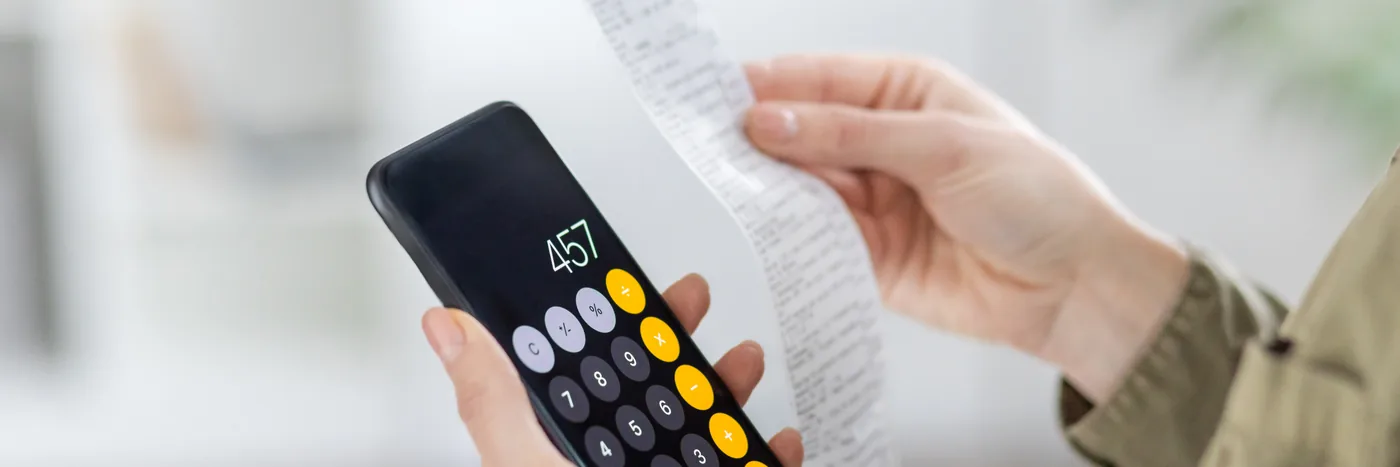A hand holding a calculator against a paper receipt while working out a household cost.