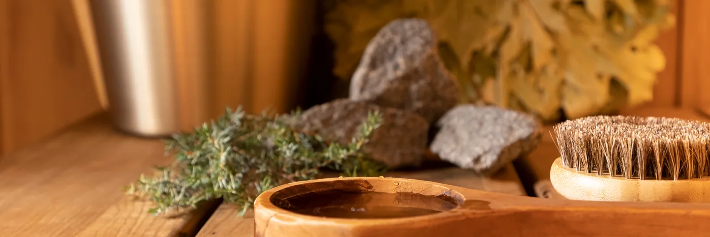 Sauna accessories on a wooden bench: a birch ladle, sauna stones, dried herbs and a bristle brush.