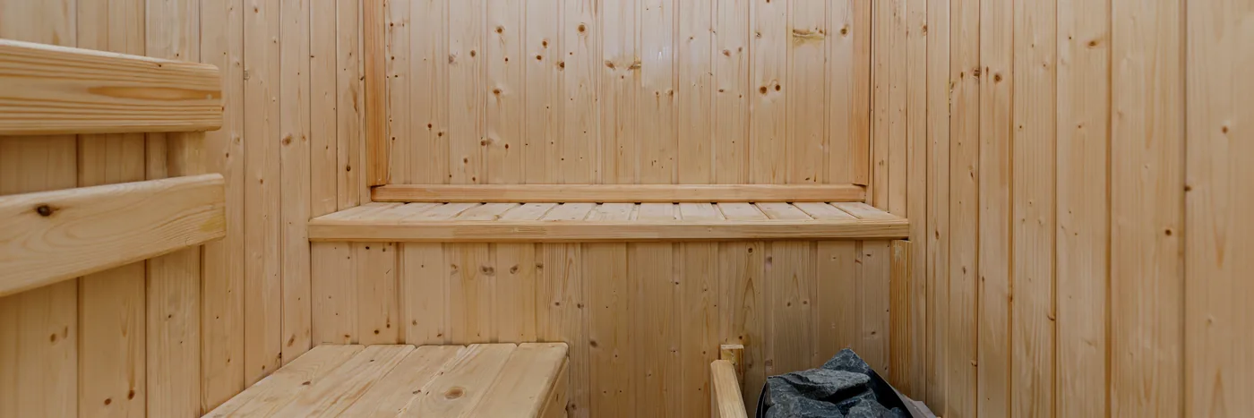 The pale wooden interior of a small home sauna room, with a bench and a heater in shadow.
